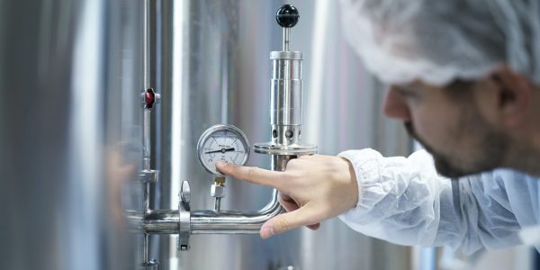 Technologist in white protective suit checking pressure on pressure gauge on industrial machine in factory.